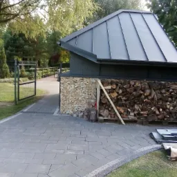 Nowoczesny budynek gospodarczy z szarym dachem panelowym, ścianą z piaskowca i stosem drewna na palecie, widok z brukowanego podjazdu w kierunku otwartej bramy.