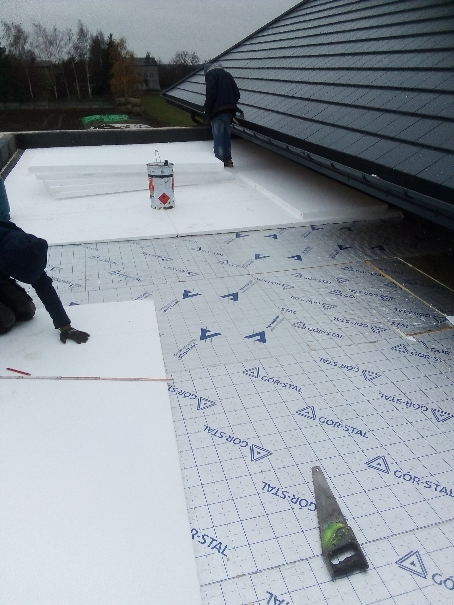 Prace dekarskie: układanie izolacji termicznej z białych płyt styropianowych na membranie GOR-STAL na płaskim dachu, widoczna piła ręczna i puszka z klejem.