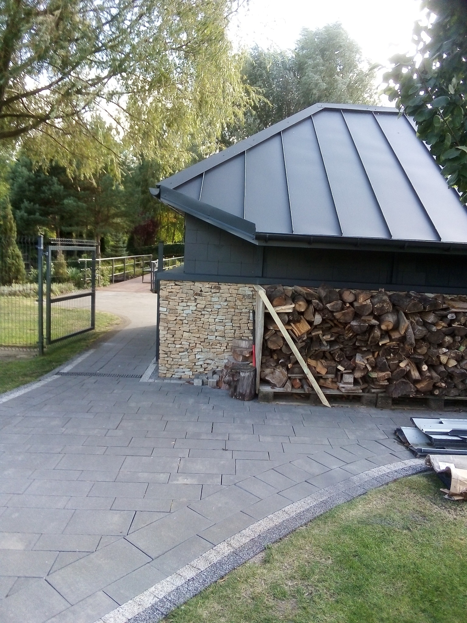 Nowoczesny budynek gospodarczy z szarym dachem panelowym, ścianą z piaskowca i stosem drewna na palecie, widok z brukowanego podjazdu w kierunku otwartej bramy.