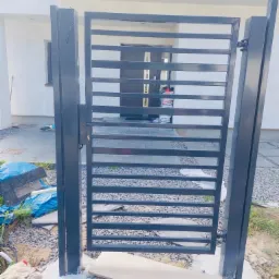 Czarna, metalowa brama wjazdowa z poziomymi szczeblami, zamontowana przed nowoczesnym domem z szarą stolarką okienną i drzwiami wejściowymi, widoczne ślady prac montażowych wokół fundamentów bramy.