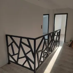 Czarna, metalowa balustrada o geometrycznym wzorze, zamontowana na antresoli z podłogą imitującą drewno, widok na białe ściany i drzwi w ciemnych futrynach.