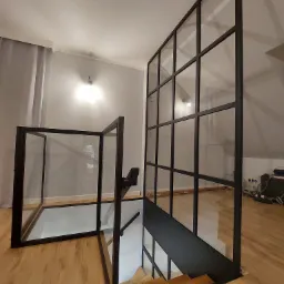 Nowoczesne schody z drewnianymi stopniami zabezpieczone czarną, metalową konstrukcją z przezroczystymi panelami szklanymi, tworzącymi balustradę i ściankę działową.