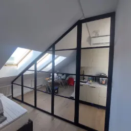 Przeszklona ścianka działowa z czarnymi szprosami oddzielająca sypialnię na poddaszu od kuchni, widok na okno dachowe i grzejnik.