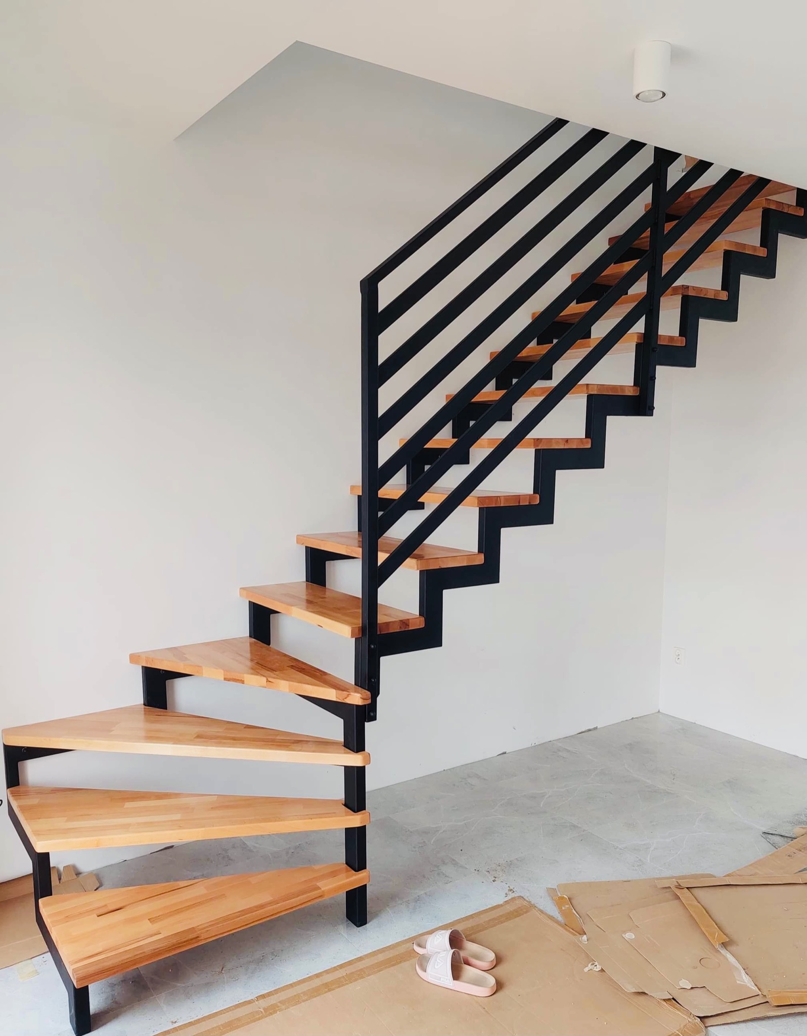 Drewniane schody z czarną, metalową balustradą w nowoczesnym wnętrzu, widoczne kartony i obuwie na podłodze.