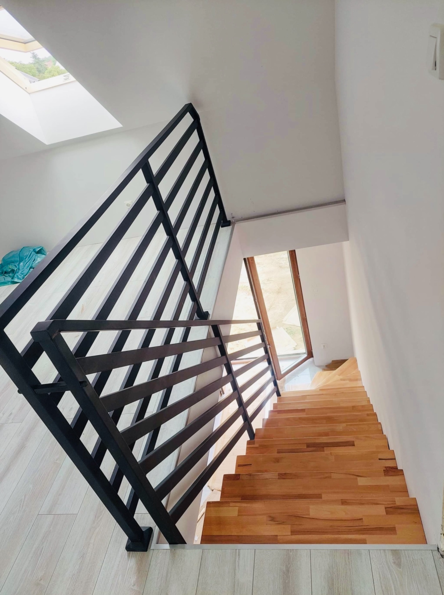Drewniane schody z czarną, nowoczesną balustradą z metalowych profili, widok z góry, jasne wnętrze z oknem dachowym.