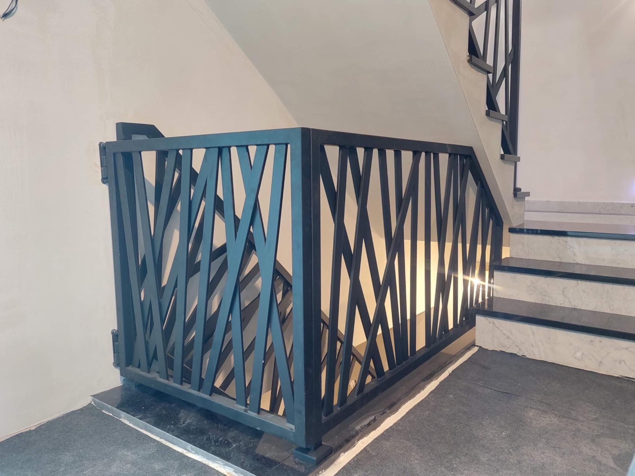 Metalowa balustrada schodowa z geometrycznym wzorem krzyżujących się prętów, pomalowana na ciemny kolor, zamontowana na marmurowych schodach z czarnymi akcentami.