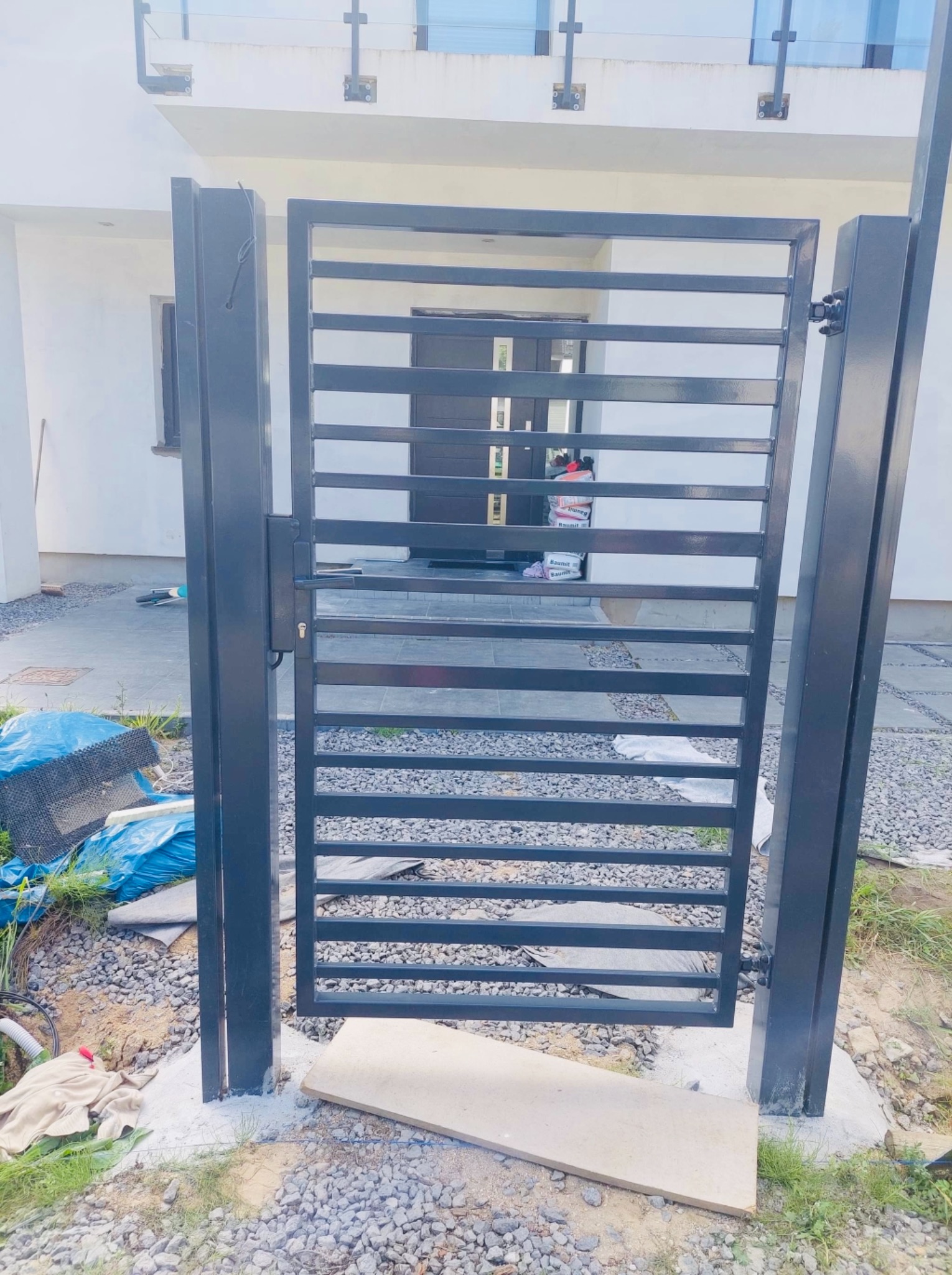 Czarna, metalowa brama wjazdowa z poziomymi szczeblami, zamontowana przed nowoczesnym domem z szarą stolarką okienną i drzwiami wejściowymi, widoczne ślady prac montażowych wokół fundamentów bramy.