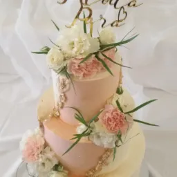 Trzypiętrowy tort weselny w odcieniach różu i kremu, udekorowany świeżymi kwiatami i złotym napisem 'Młoda Para', na tle białej tkaniny.