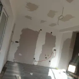 Wnętrze w trakcie remontu: częściowo położone szare płytki podłogowe, ściany z widocznymi śladami szpachlowania i instalacji elektrycznej, sufit z miejscowymi poprawkami gipsowymi.