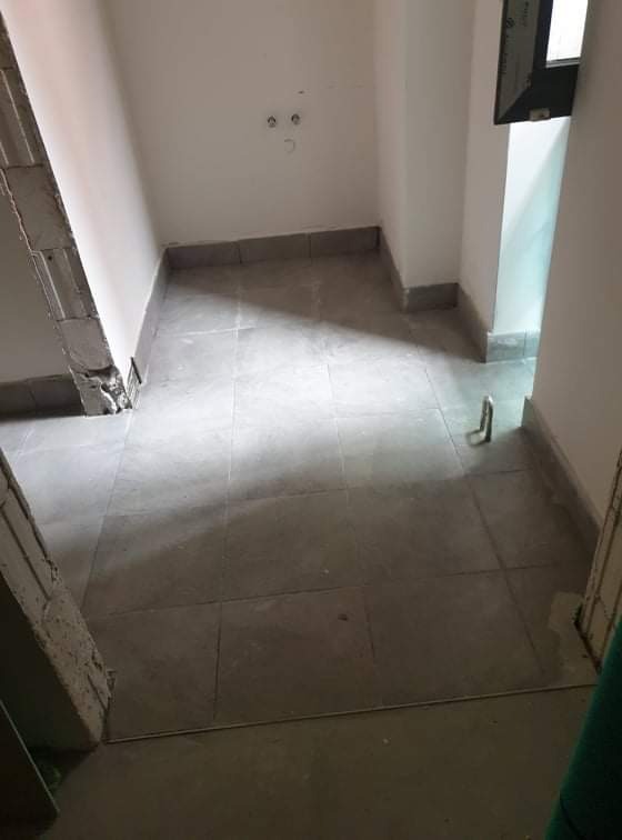 Fragment wnętrza w trakcie remontu: szare płytki podłogowe, surowe ściany z widocznymi instalacjami elektrycznymi, obróbka przy podłodze z płytek, widoczny fragment niedokończonego muru w rogu...