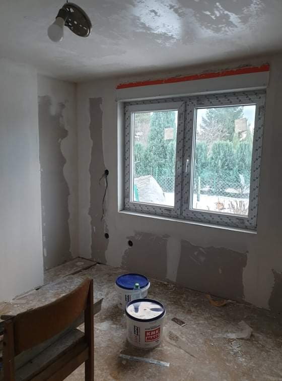 Pokój w trakcie prac wykończeniowych: ściany częściowo pokryte gładzią, dwa wiadra z masą szpachlową na podłodze, widoczne instalacje elektryczne, okno z widokiem na ogród.