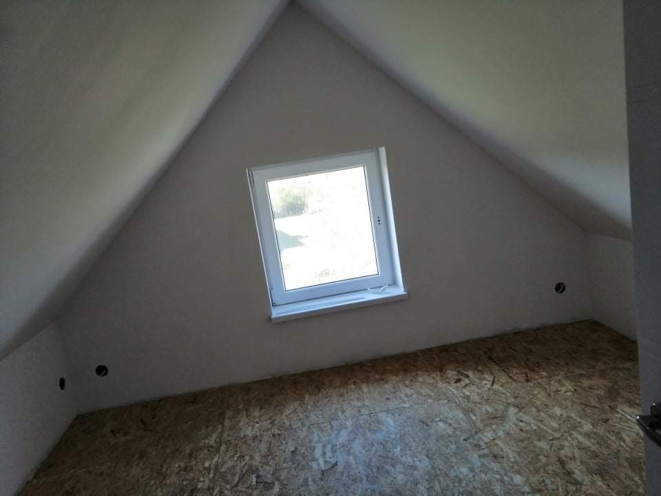 Poddasze z oknem, surowa płyta OSB na podłodze, ściany przygotowane do malowania, widoczne otwory na gniazdka elektryczne.