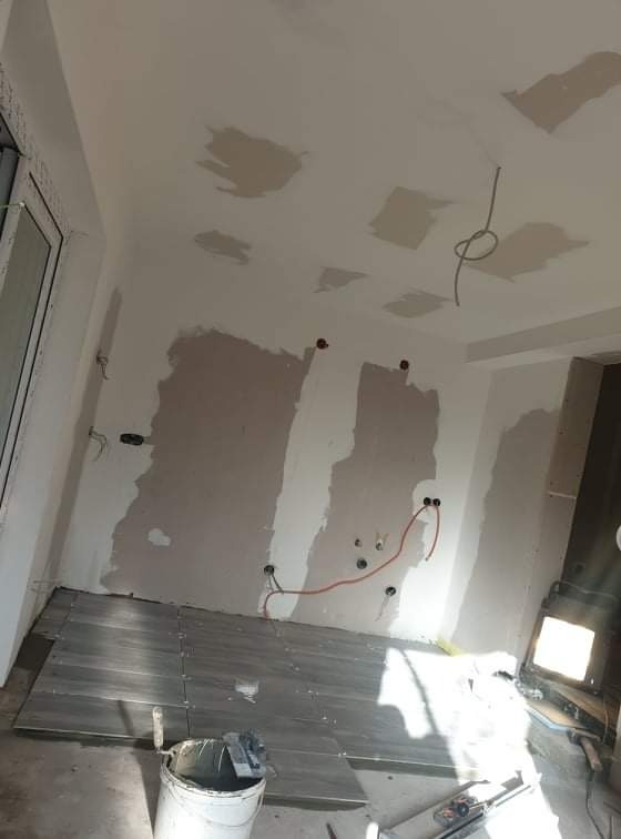 Wnętrze w trakcie remontu: częściowo położone szare płytki podłogowe, ściany z widocznymi śladami szpachlowania i instalacji elektrycznej, sufit z miejscowymi poprawkami gipsowymi.