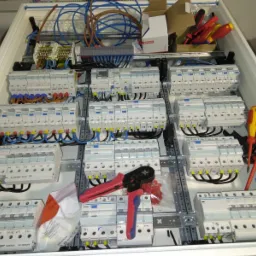 Rozdzielnica elektryczna w trakcie montażu z widocznymi bezpiecznikami, przewodami w kolorach niebieskim, żółtym i czarnym, narzędziami oraz elementami montażowymi.