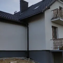 Świeżo otynkowany dom z ciemną podmurówką i brązową stolarką okienną, widoczne rusztowanie przy ścianie szczytowej.