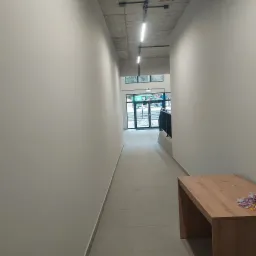 Korytarz w budynku po remoncie, z jasnymi ścianami, betonowym sufitem z oświetleniem szynowym i drewnianym stolikiem z ulotkami w rogu.