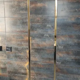 Ściana wyłożona płytkami imitującymi drewno w odcieniach brązu i szarości, z pionowymi, złotymi listwami dekoracyjnymi, oraz fragment podłogi z płytek w podobnej kolorystyce.