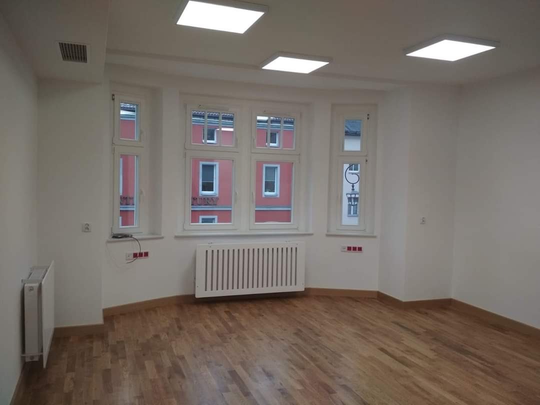 Puste, jasne pomieszczenie z parkietem, białymi ścianami, trzema oknami z białymi ramami, grzejnikami i kwadratowymi lampami sufitowymi.