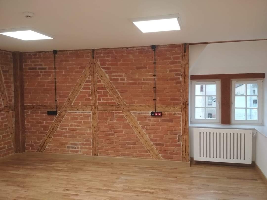 Wnętrze pokoju z odsłoniętą ceglaną ścianą z drewnianą konstrukcją, parkietem na podłodze, białym grzejnikiem pod dwoma oknami i nowoczesnym oświetleniem sufitowym.