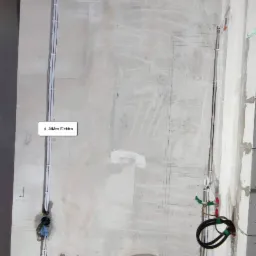 Surowa ściana z widocznymi przewodami elektrycznymi i instalacjami wodno-kanalizacyjnymi w trakcie montażu, z zaznaczeniami i notatkami na ścianie.