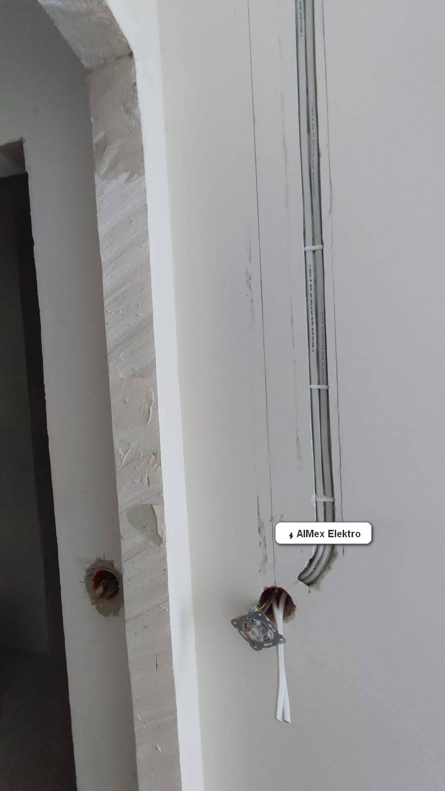 Świeżo poprowadzone kable w białej rurce PCV, przytwierdzone do ściany klamrami, oraz puszka pod gniazdko z wystającymi przewodami, widoczne ślady po kuciu w ścianie.