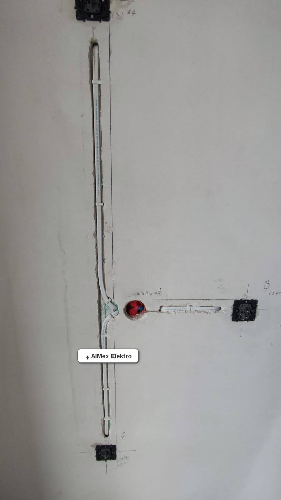 Ściana w trakcie remontu z widocznymi przewodami elektrycznymi w białych rurkach, puszkami pod gniazdka i napisami odręcznymi na ścianie.