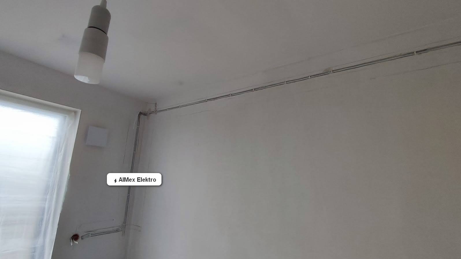 Białe kable elektryczne poprowadzone po ścianie i suficie w nowym mieszkaniu, widoczne puszki instalacyjne, lampa sufitowa i okno zasłonięte folią malarską.