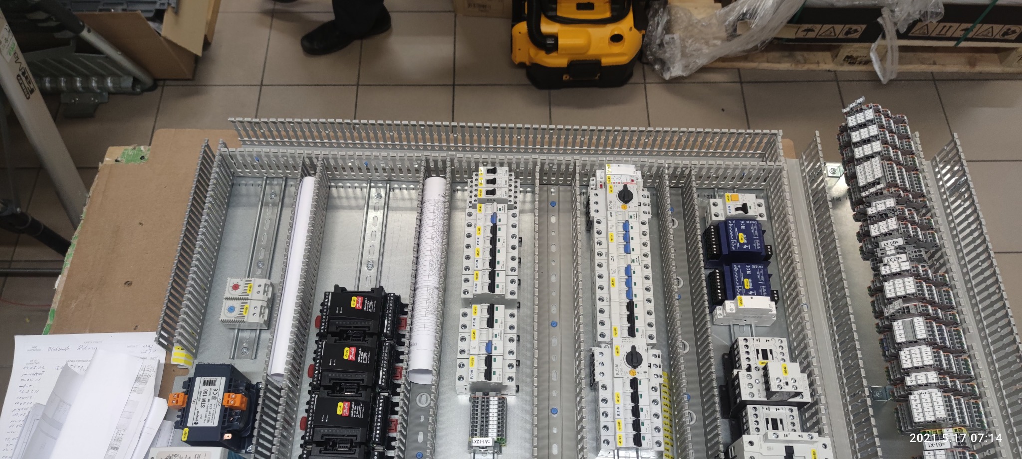 Widok z góry na rozdzielnicę elektryczną w trakcie montażu, widoczne liczne moduły, przekaźniki, bezpieczniki i dokumentacja techniczna, częściowo zamontowane na szynach DIN.