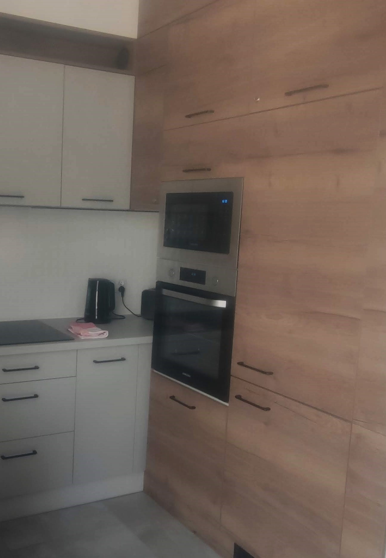 Nowoczesna kuchnia z zabudową meblową w odcieniach szarości i drewna, z wbudowanym piekarnikiem i płytą indukcyjną oraz czajnikiem elektrycznym na blacie.