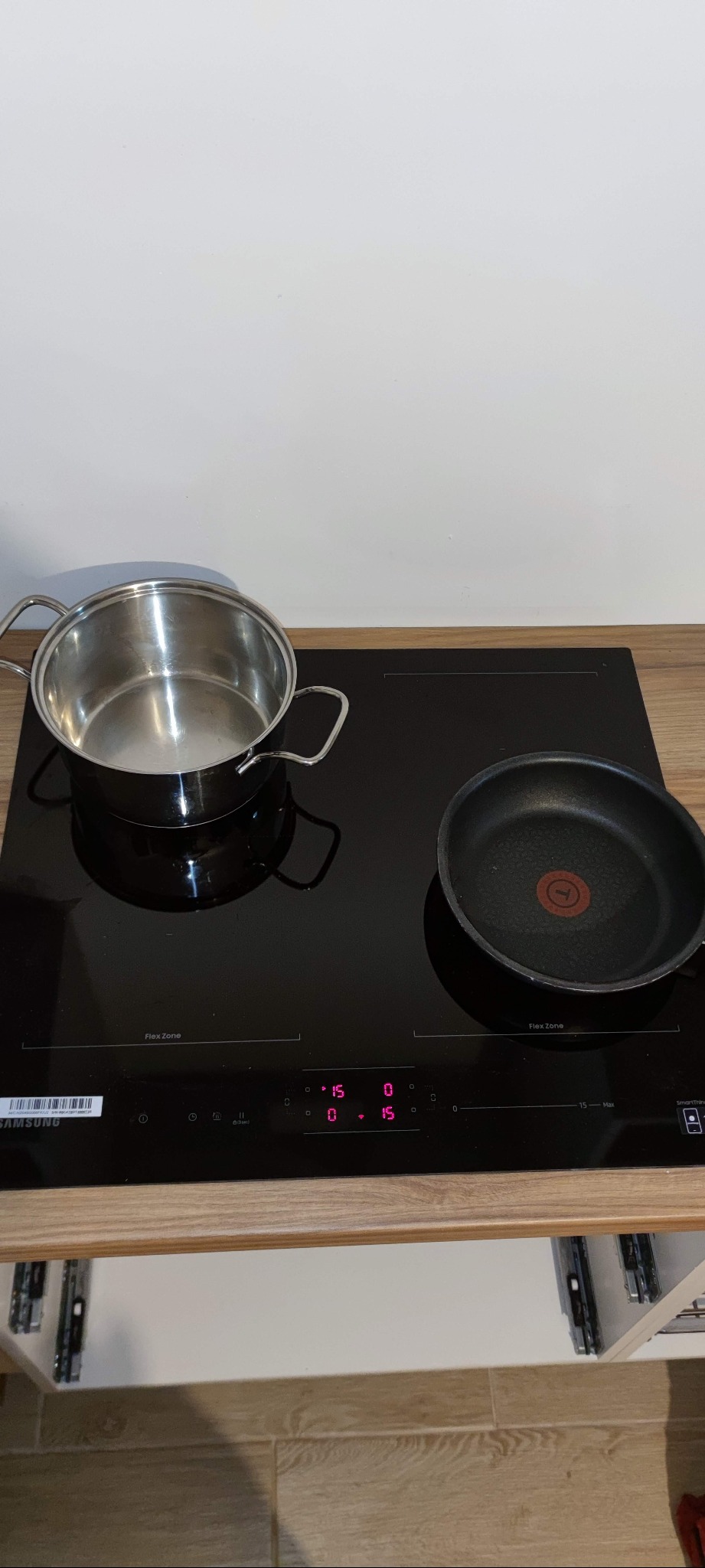 Czarna płyta indukcyjna Samsung z włączonymi polami grzewczymi, na których stoją garnek ze stali nierdzewnej i patelnia z czerwonym punktem na środku, ustawiona na blacie kuchennym z drewnopodobnym...