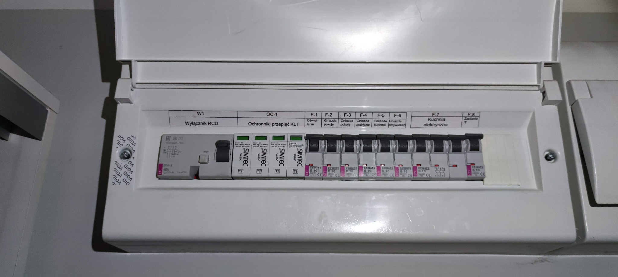 Otwarta skrzynka elektryczna z wyłącznikiem RCD, ochronnikami przepięć i bezpiecznikami oznaczonymi dla różnych obwodów: oświetlenie, gniazda pokojowe, pralka/łazienka, kuchnia, zmywarka/okap...