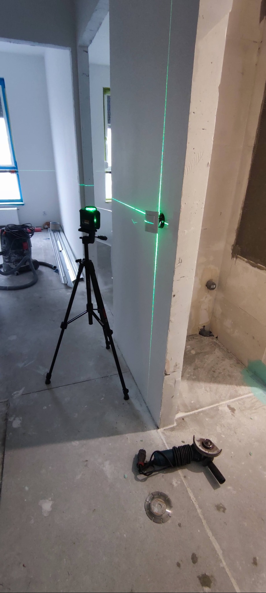 Poziomica laserowa ustawiona na trójnogu, wyznaczająca pion na ścianie z zamontowanym gniazdkiem elektrycznym w surowym wnętrzu, obok leży szlifierka kątowa i tarcza diamentowa.