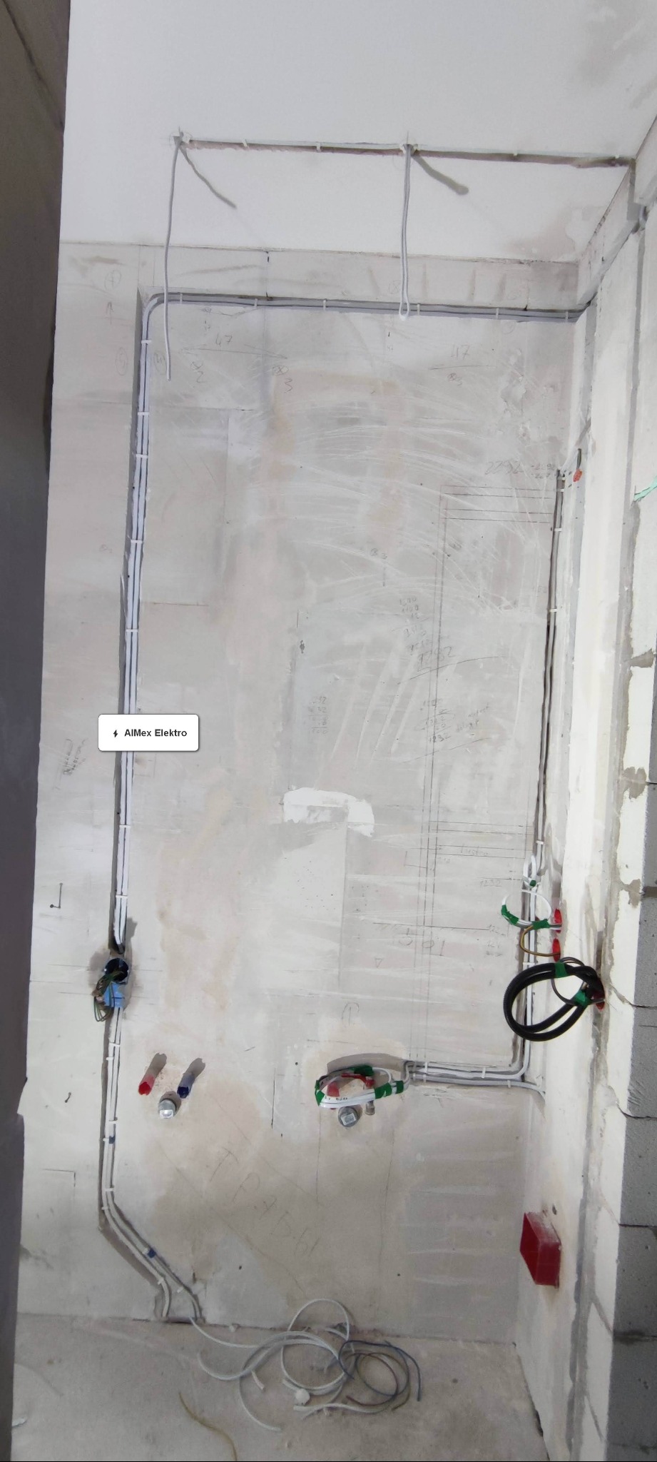 Surowa ściana z widocznymi przewodami elektrycznymi i instalacjami wodno-kanalizacyjnymi w trakcie montażu, z zaznaczeniami i notatkami na ścianie.