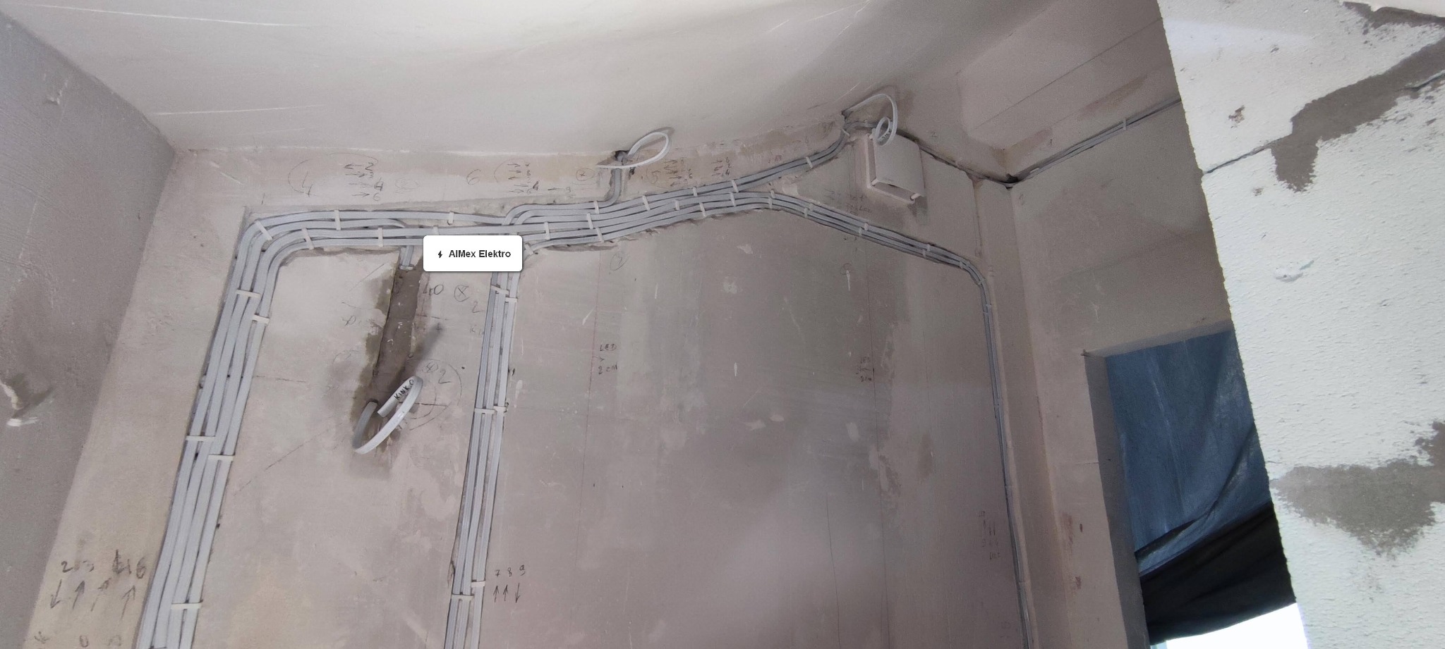 Szereg białych kabli elektrycznych przymocowanych do surowej ściany w trakcie instalacji, widoczne oznaczenia i notatki na ścianie, fragment otworu drzwiowego z zasłoniętym wejściem.
