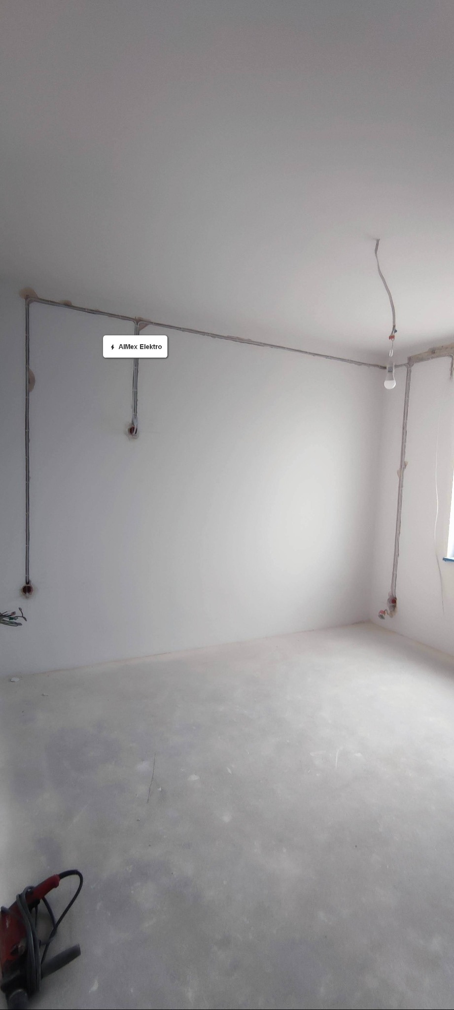 Puste pomieszczenie w trakcie instalacji elektrycznej, widoczne kable poprowadzone wzdłuż ścian pod sufitem, gniazdka elektryczne i wisząca żarówka, na podłodze leży wiertarka.