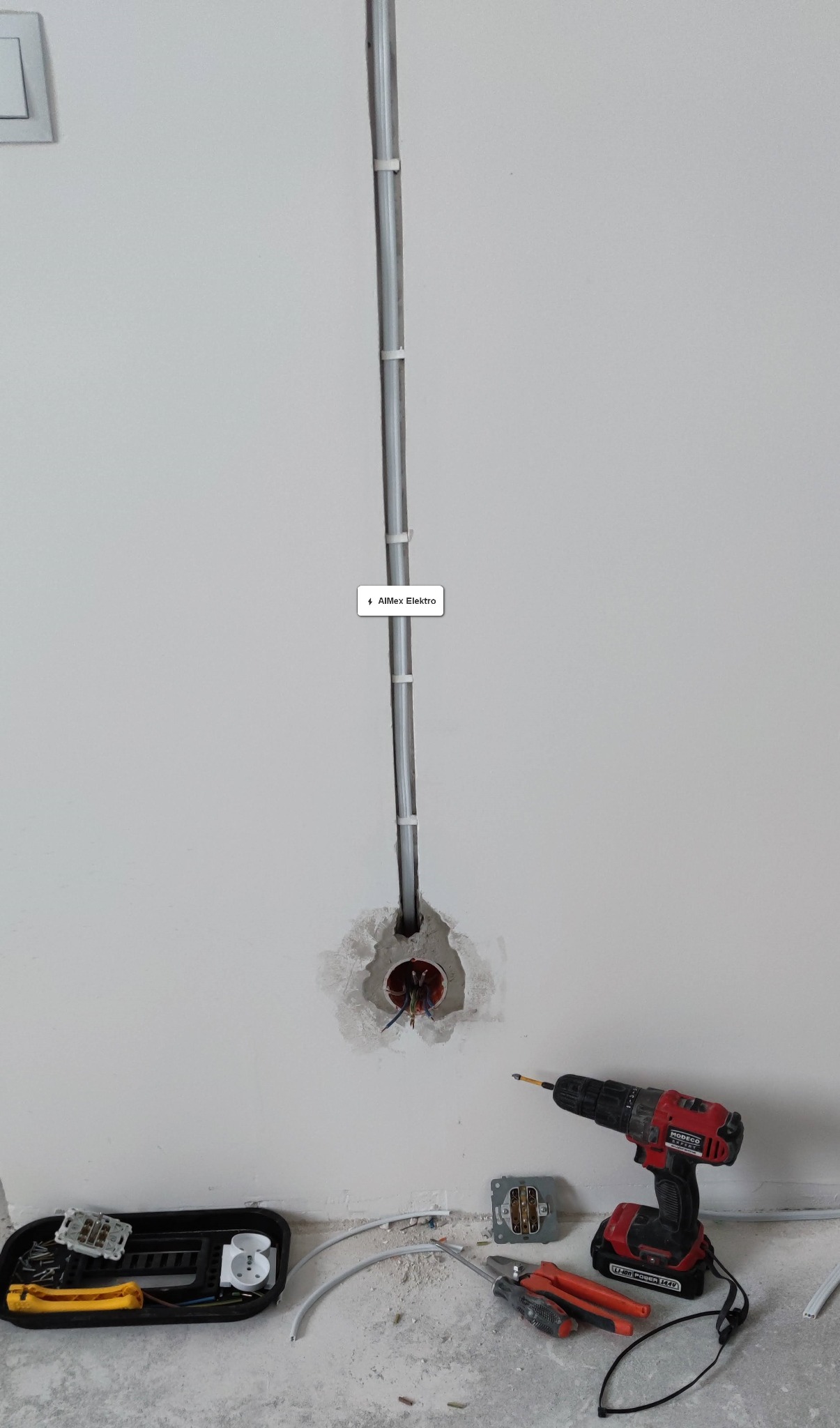Szara rura elektroinstalacyjna przymocowana do ściany z widocznymi przewodami w puszce, obok wiertarka akumulatorowa Moreco, kombinerki, nóż do zdejmowania izolacji i gniazdko elektryczne w trakcie...