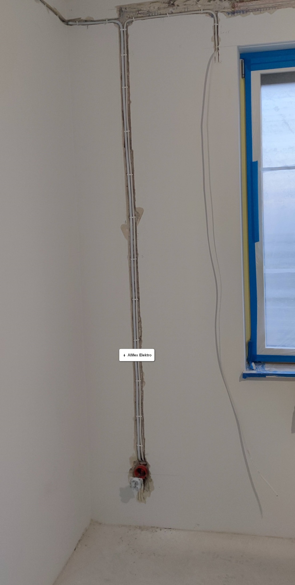 Świeżo poprowadzone białe przewody elektryczne przymocowane klamrami w bruździe ściennej, widoczne niedokończone gniazdko elektryczne z czerwonym elementem w ścianie, okno zabezpieczone taśmą...