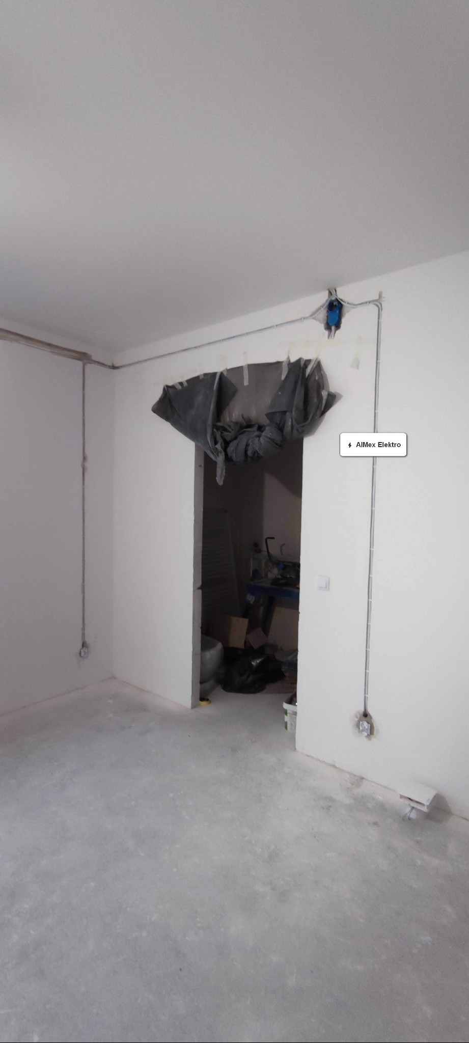 Surowe wnętrze w trakcie remontu, widoczne poprowadzone kable elektryczne wzdłuż ścian i pod sufitem, zabezpieczone folią drzwi do sąsiedniego pomieszczenia.