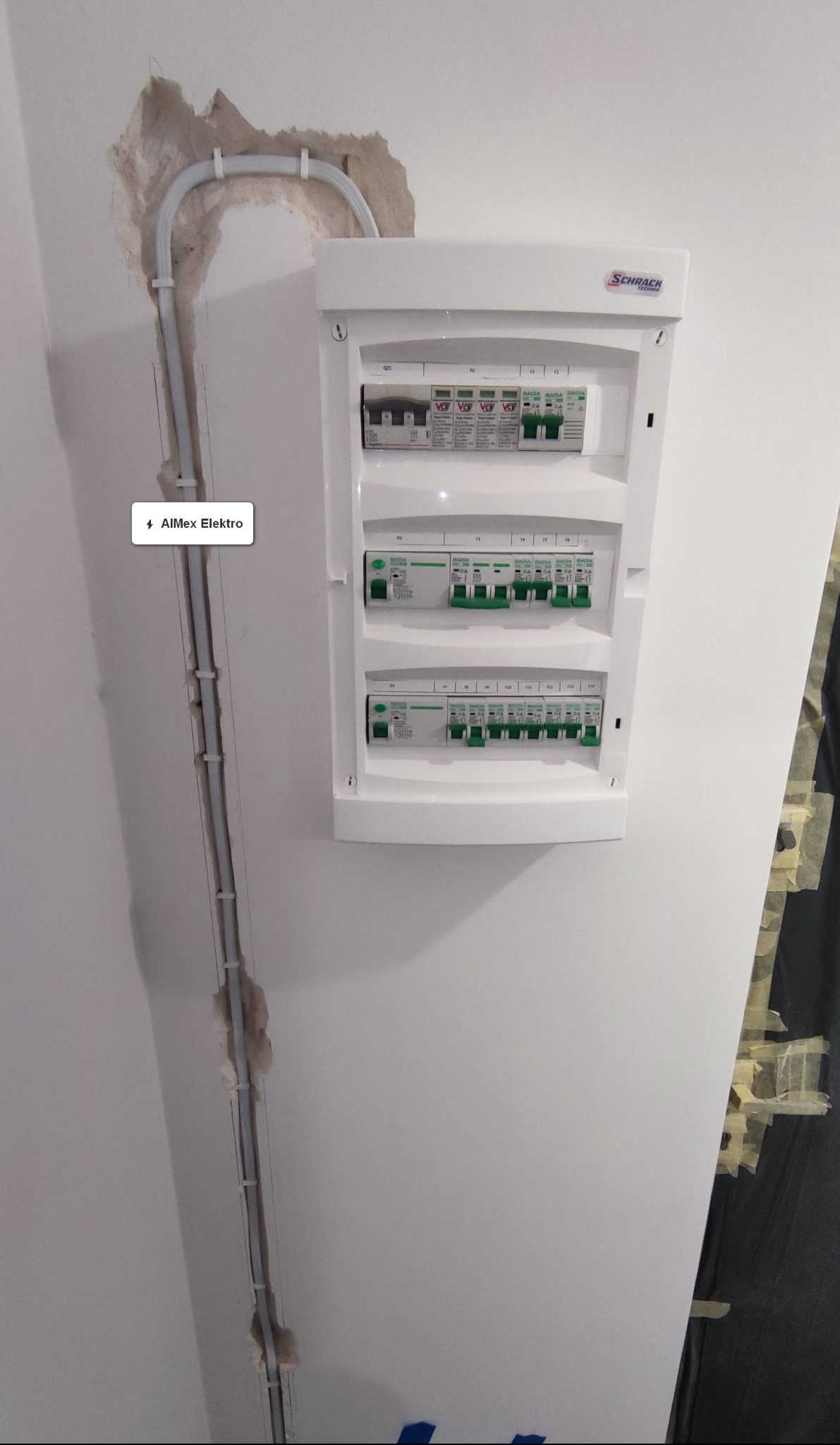 Biała skrzynka rozdzielcza firmy Schrack zamontowana na ścianie, z widocznymi zabezpieczeniami i przewodami w listwie, częściowo ukrytymi w ścianie z widocznymi ubytkami tynku.