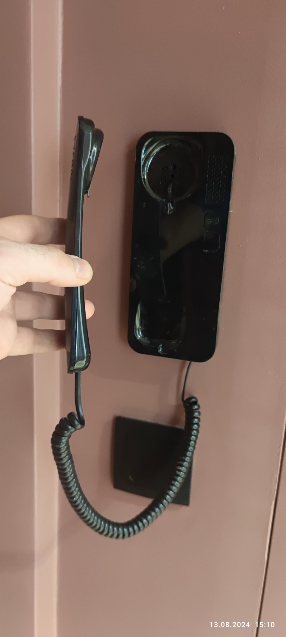 Czarny domofon z kablem spiralnym zamontowany na różowej ścianie, trzymany w dłoni podczas instalacji.