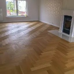 Jasny parkiet ułożony we wzór jodełki w przestronnym pokoju z kominkiem i oknem z widokiem na zieleń.