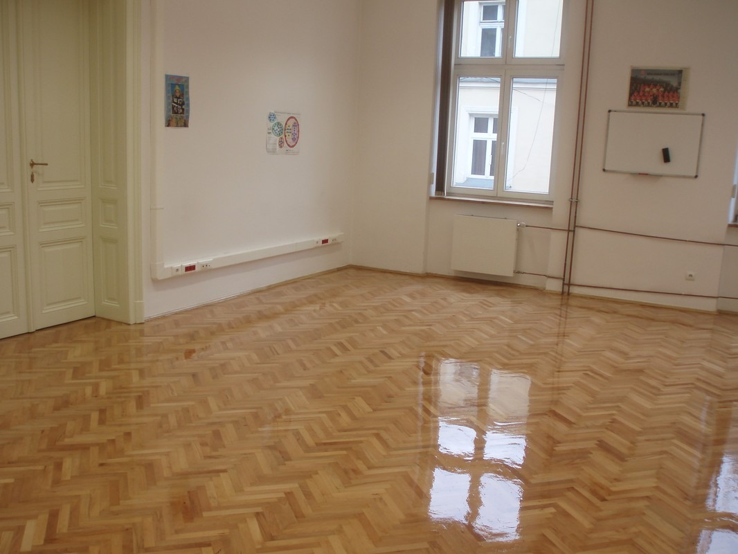 Błyszcząca, odnowiona podłoga parkietowa ułożona w jodełkę, odbijająca światło okna w pustym, jasnym pokoju z białą tablicą i grzejnikiem.