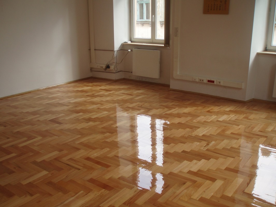 Świeżo polakierowany parkiet ułożony w jodełkę, z widocznym odbiciem światła w mokrej jeszcze powierzchni, w jasnym pokoju z białymi ścianami i grzejnikiem pod oknem.