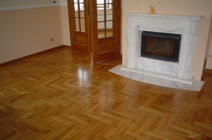 Jasny parkiet ułożony w jodełkę w przestronnym salonie z marmurowym kominkiem i drewnianymi drzwiami balkonowymi.