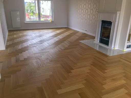 Jasny parkiet ułożony we wzór jodełki w przestronnym pokoju z kominkiem i oknem z widokiem na zieleń.