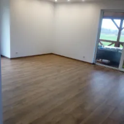 Pusty pokój z drewnianą podłogą, świeżo pomalowanymi białymi ścianami i sufitowymi lampami punktowymi, widok na zewnątrz przez duże okno balkonowe.