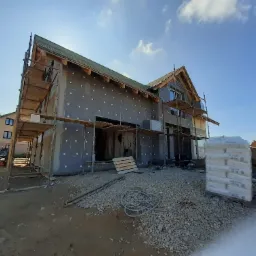 Nowo budowany dom murowany z ocieploną elewacją w trakcie prac wykończeniowych, widoczne rusztowania, styropianowe kołki mocujące, worki z materiałami budowlanymi i niebieski wąż ogrodowy...