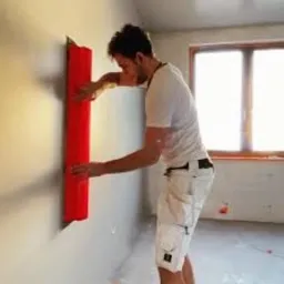 Mężczyzna w białej koszulce i kr&oacute;tkich spodenkach przykłada czerwoną rolkę do szarej, surowej ściany w jasnym, pustym pomieszczeniu. Widoczne ślady po instalacji elektrycznej.