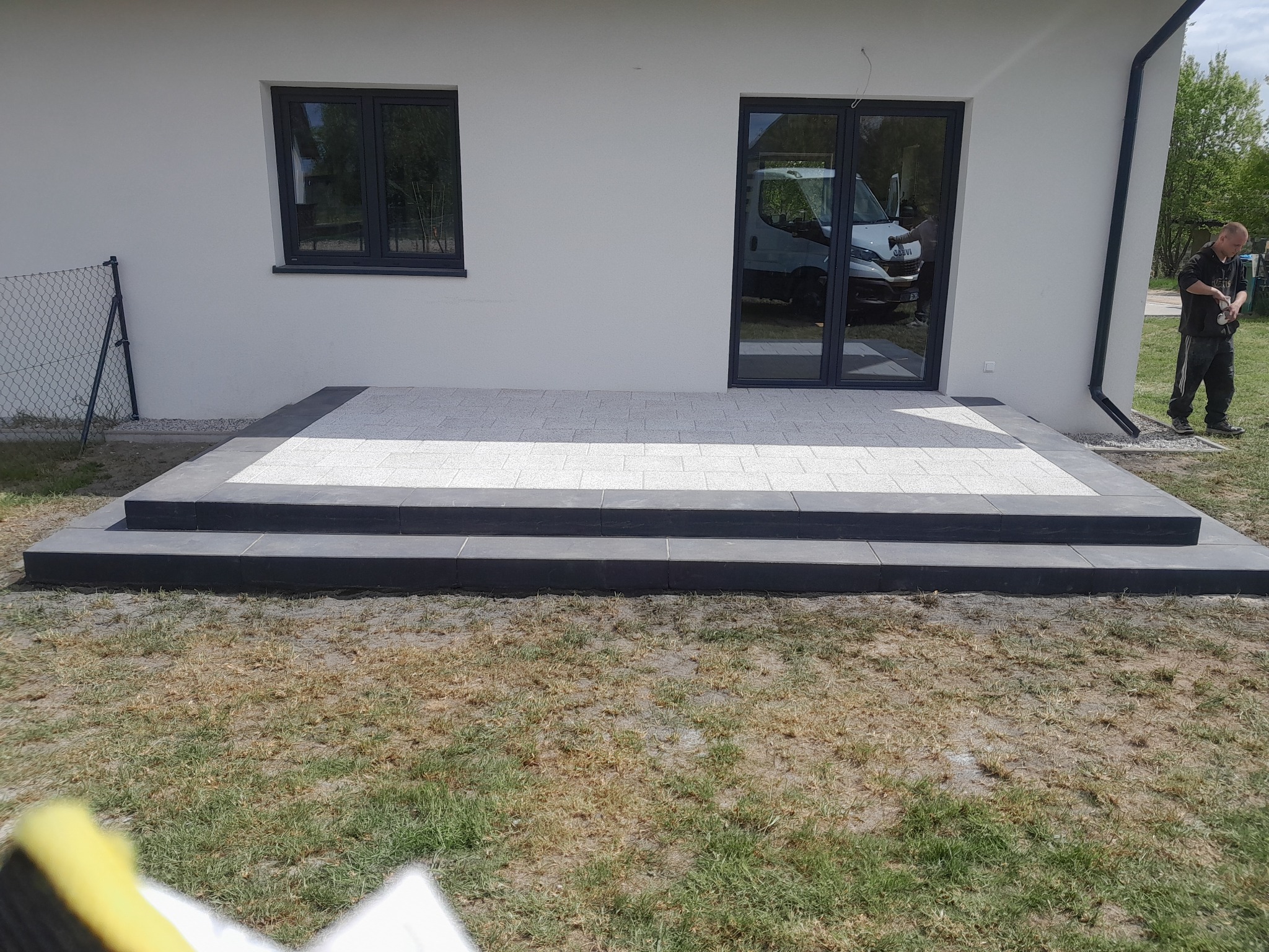 Wykończony taras z jasnej kostki brukowej przed domem z białymi ścianami i czarną stolarką okienną. Widoczne dwustopniowe schody z ciemnej kostki.