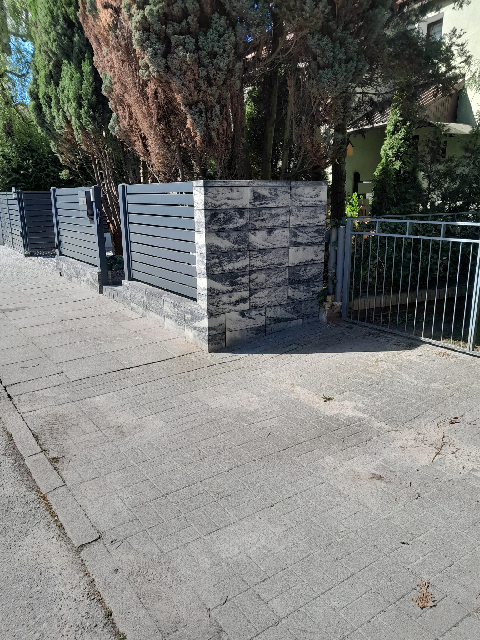 Nowoczesne ogrodzenie panelowe z grafitowymi przęsłami i słupkami z dekoracyjnej kostki, widok z perspektywy chodnika z szarej kostki brukowej.